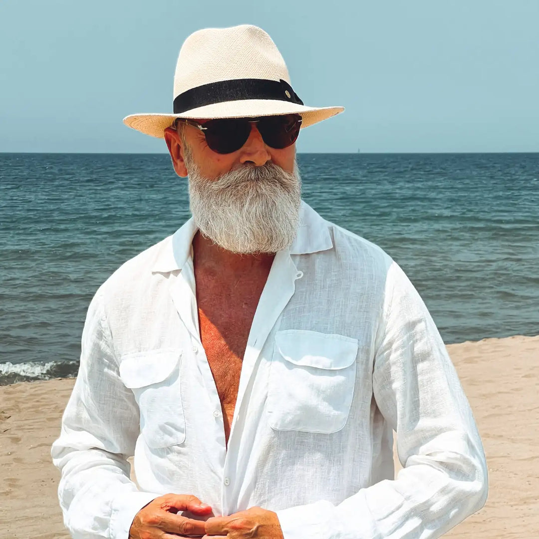 mezzo busto con sguardo a destra di un signore adulto che ricorda un gentleman di altri tempi, al mare, con barba lunga bianca, occhiali da sole e camicia bianca di lino che indossa il cappello di panama con vista quasi frontale in stile fedora color naturale di Primario Nesti