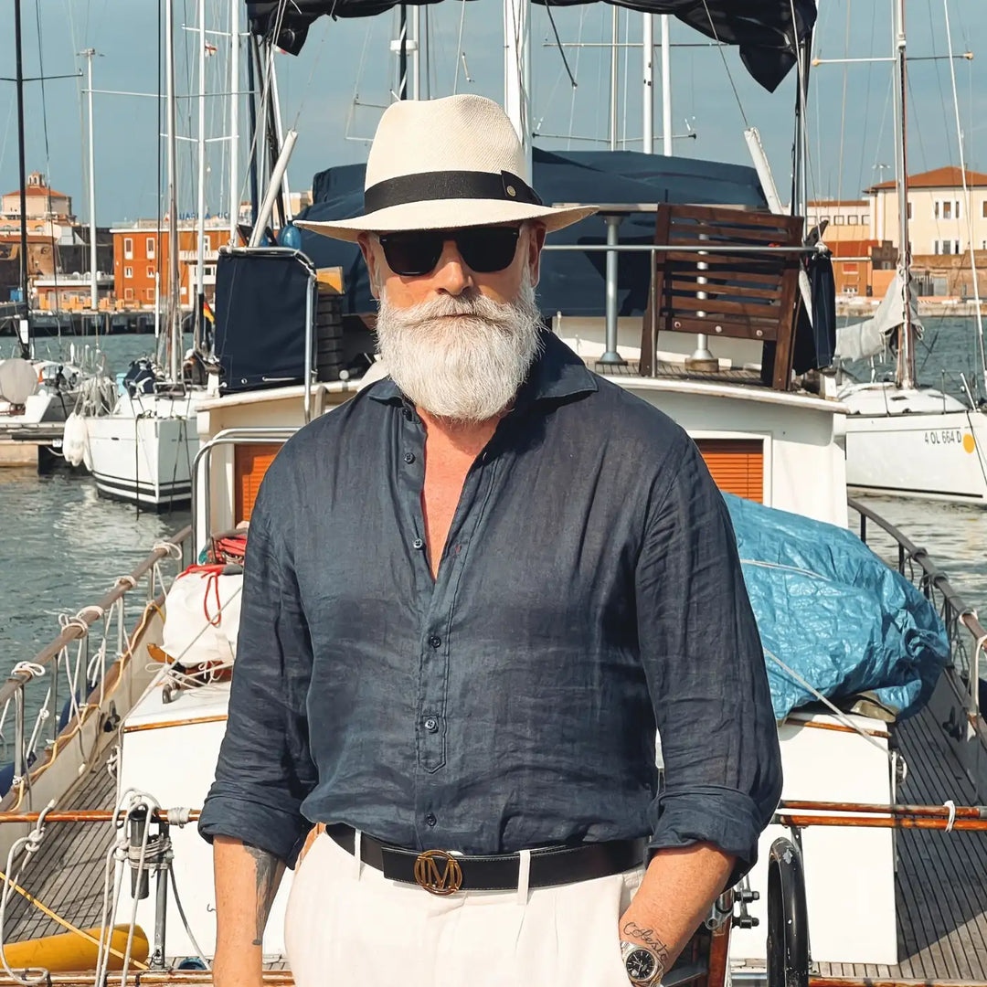 primo piano frontale di un signore adulto che ricorda un gentleman di altri tempi, al porto, con barba lunga bianca, occhiali da sole, camicia blu di lino e pantaloni bianchi, che indossa il cappello di panama in stile fedora color naturale di Primario Nesti