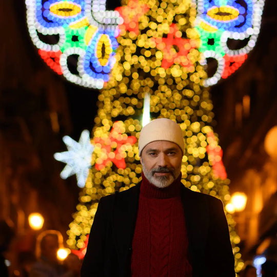 modello maturo con barba bianca indossa un berretto invernale in lana con risvolto a costine colore panna con sfondo di albero e luci natalizie